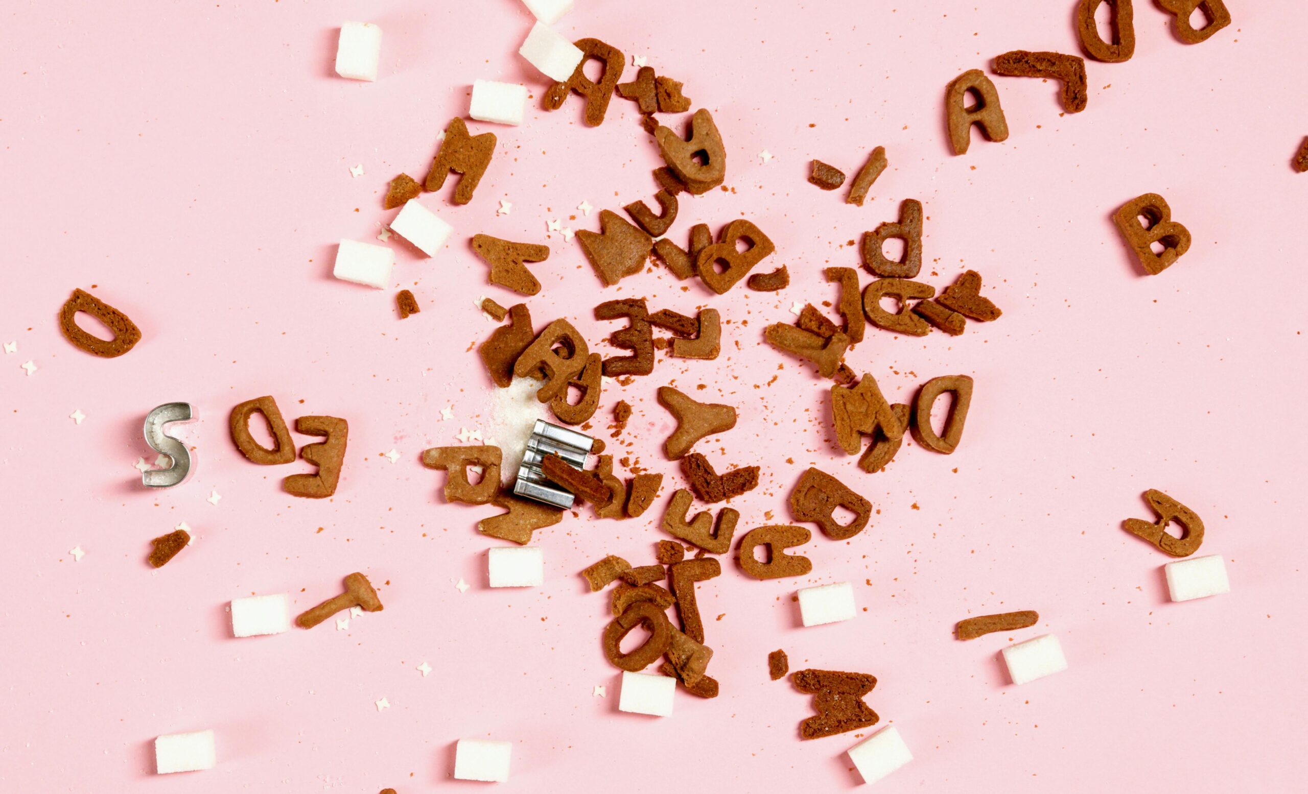 Overhead view of sugar cubes and cookie letters scattered on a pink surface, suggesting sweet chaos.