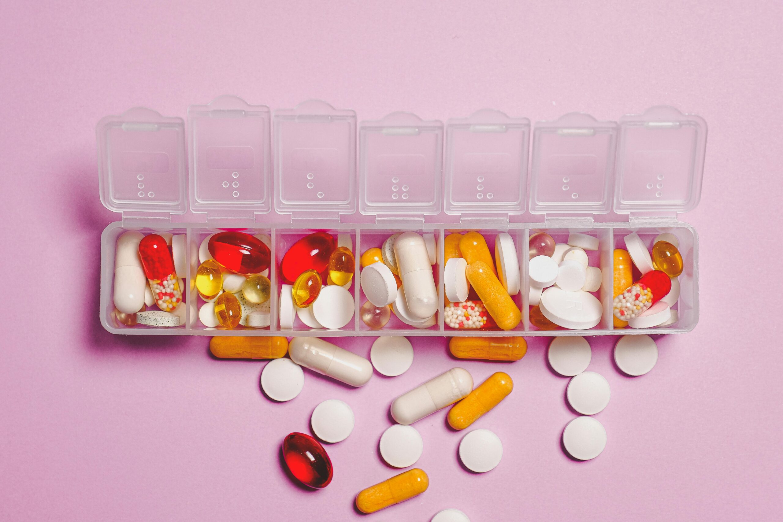 A variety of pills in a weekly pill organizer on a pink background.