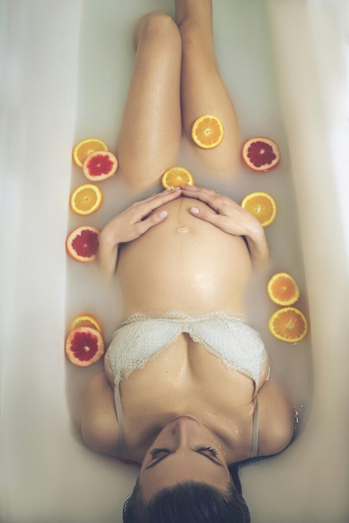 From above of calm pregnant female model in lingerie hugging big belly while taking milk bath with slices of fresh oranges and grapefruit