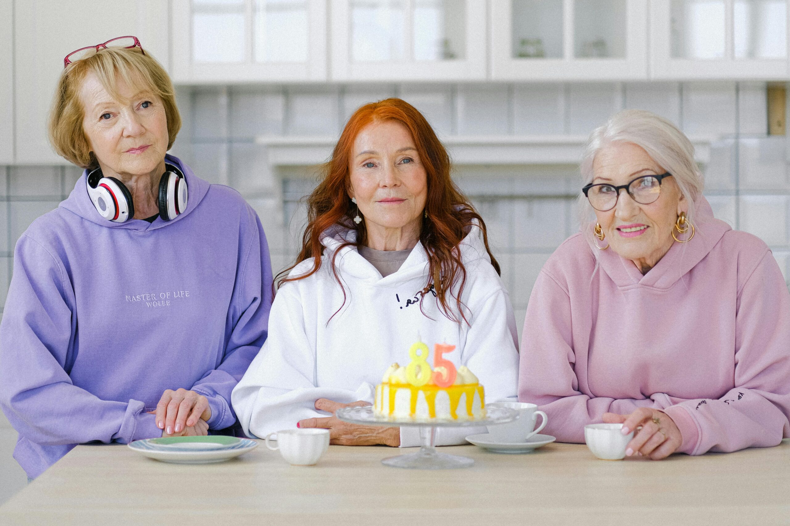 Three senior women celebrating an 85th birthday with smiles and a festive cake indoors.