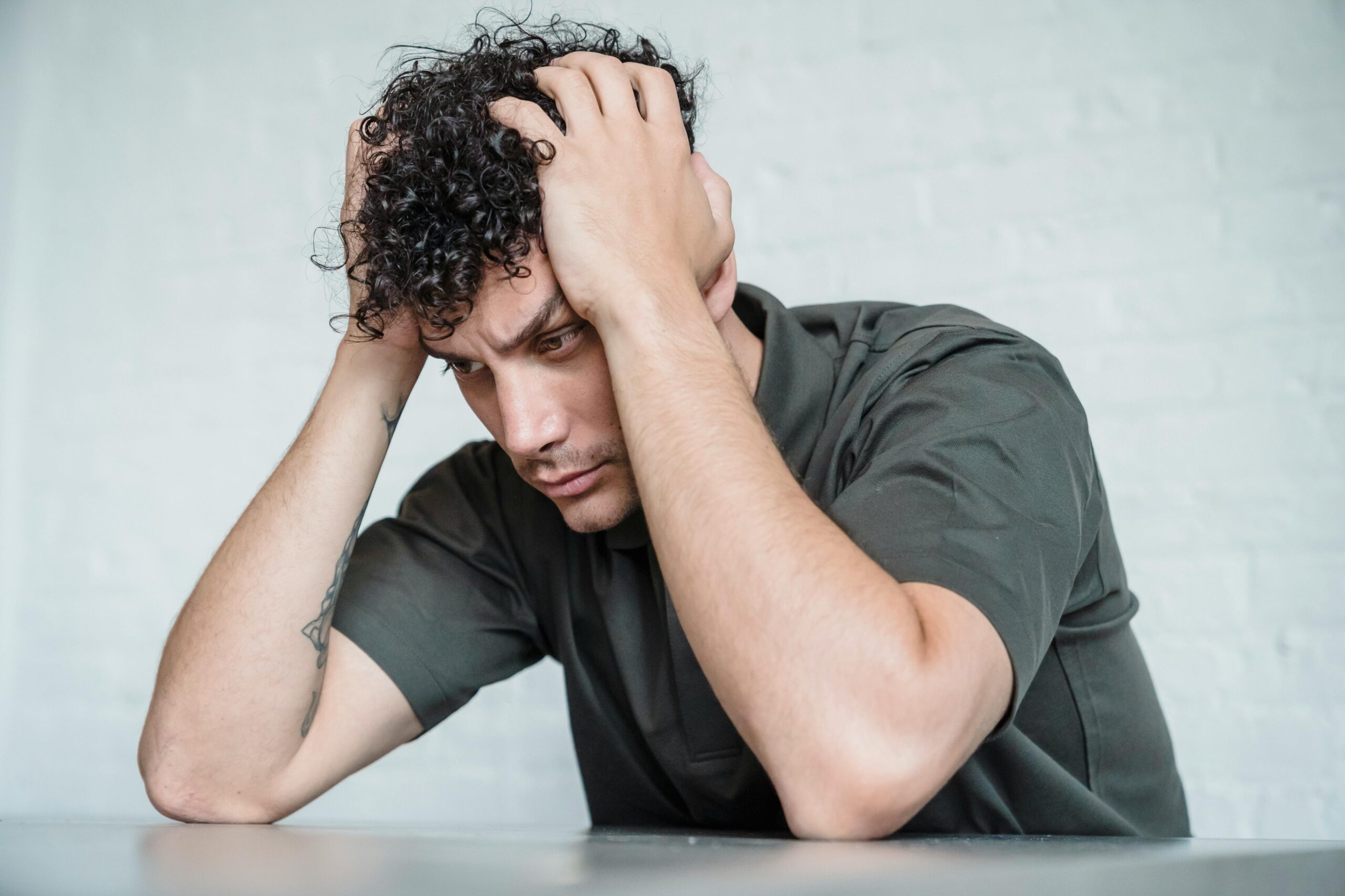 A young man with curly hair looks stressed as he sits with his hands on his head in contemplation.