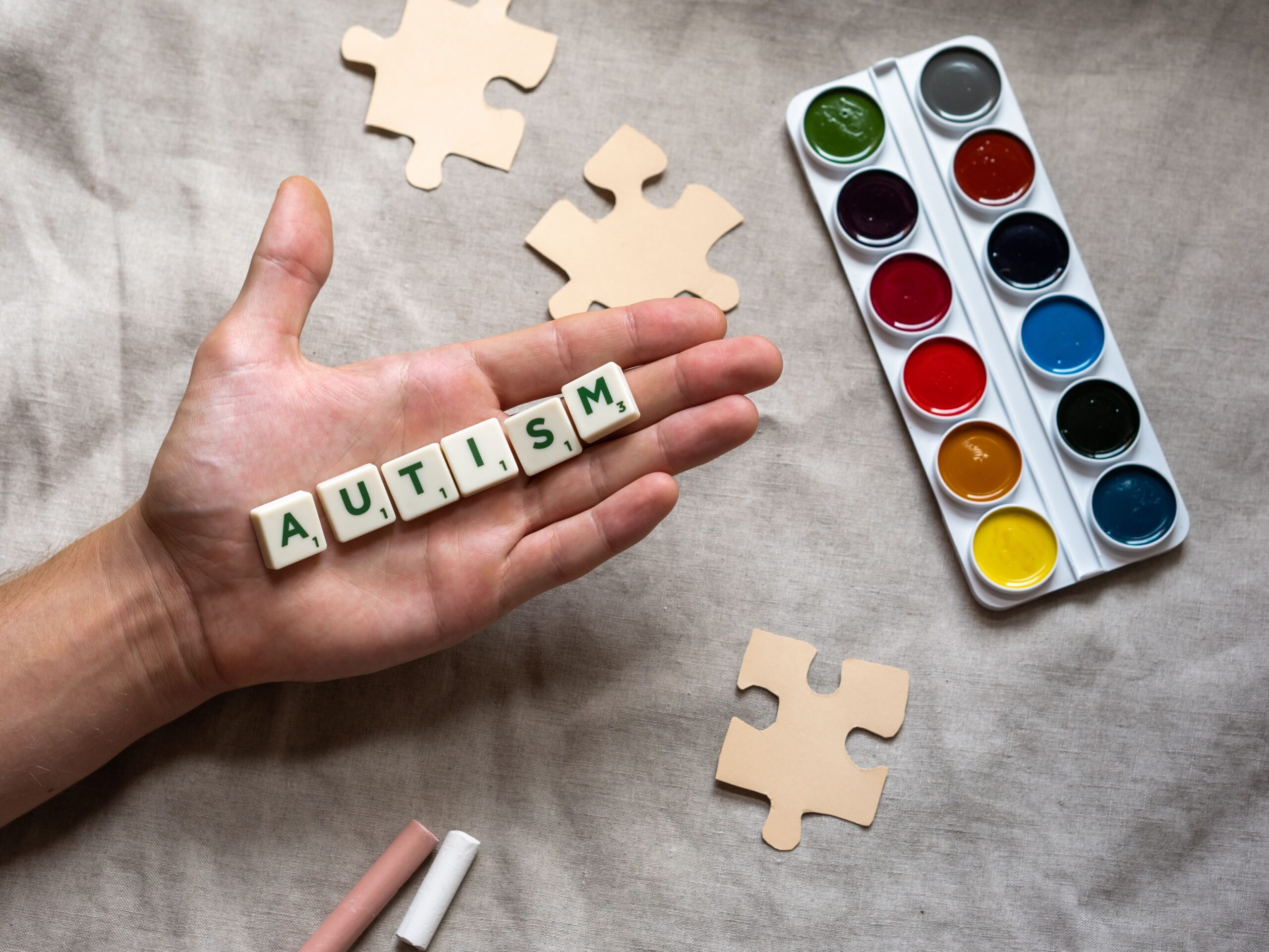 Hand holding autism scrabble tiles surrounded by puzzle pieces and watercolor paints, promoting autism awareness.